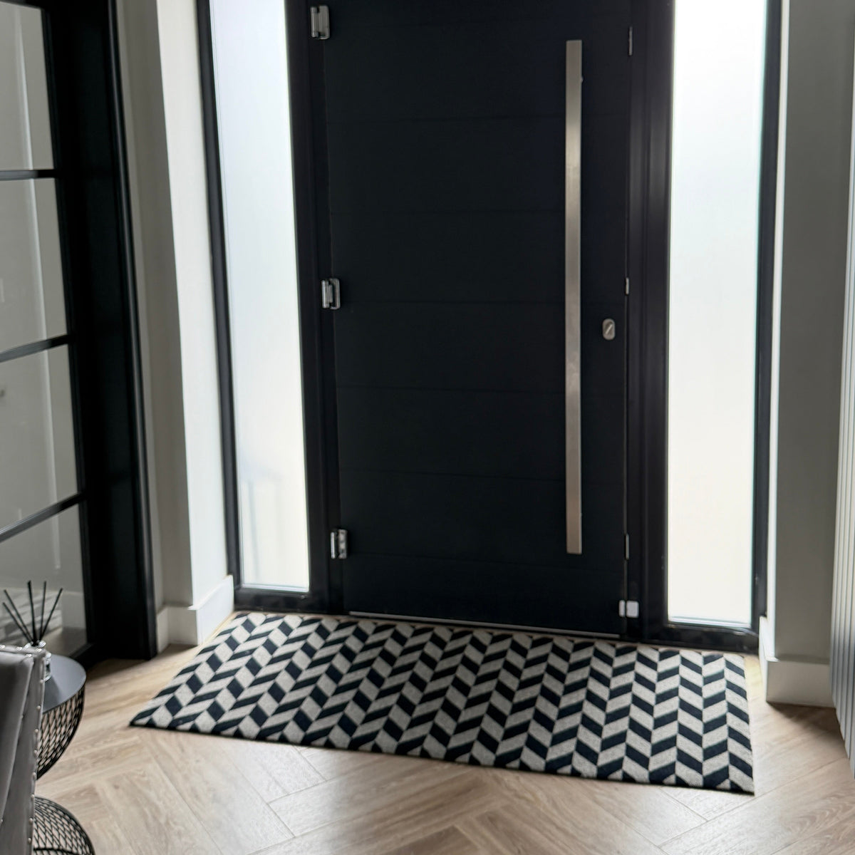 A modern black front door with a long vertical silver handle, set in a home entryway with light wood herringbone flooring, a black and white chevron-patterned doormat, and frosted side glass panels.