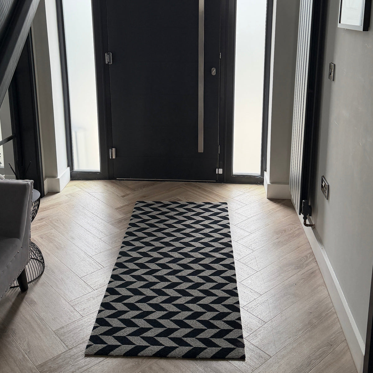 A black and white geometric patterned runner rug laid on a light wood herringbone floor in a modern entryway with a black front door and gray walls.