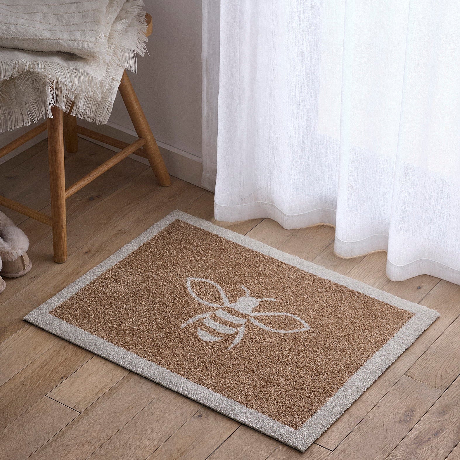 Beige doormat with a white bee design on a wooden floor next to a chair and curtains.