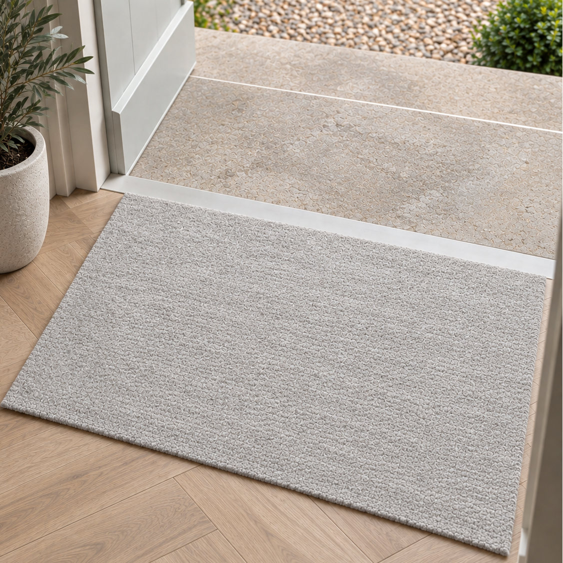 A light gray textured rectangular doormat positioned at a doorway, transitioning from a light wood herringbone floor to a concrete patio and outdoor gravel area.