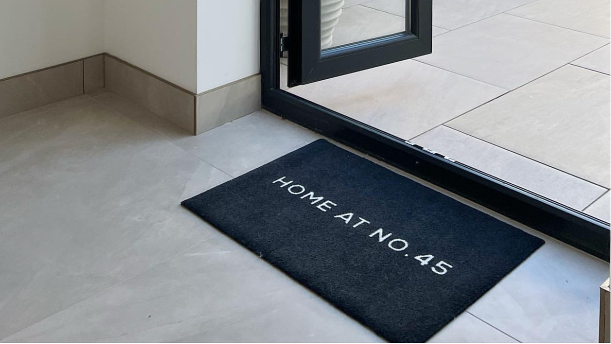 A black rectangular doormat with white text reading 'HOME AT NO. 415' placed on a light stone floor in front of an open glass patio door, with an outdoor seating area visible beyond.