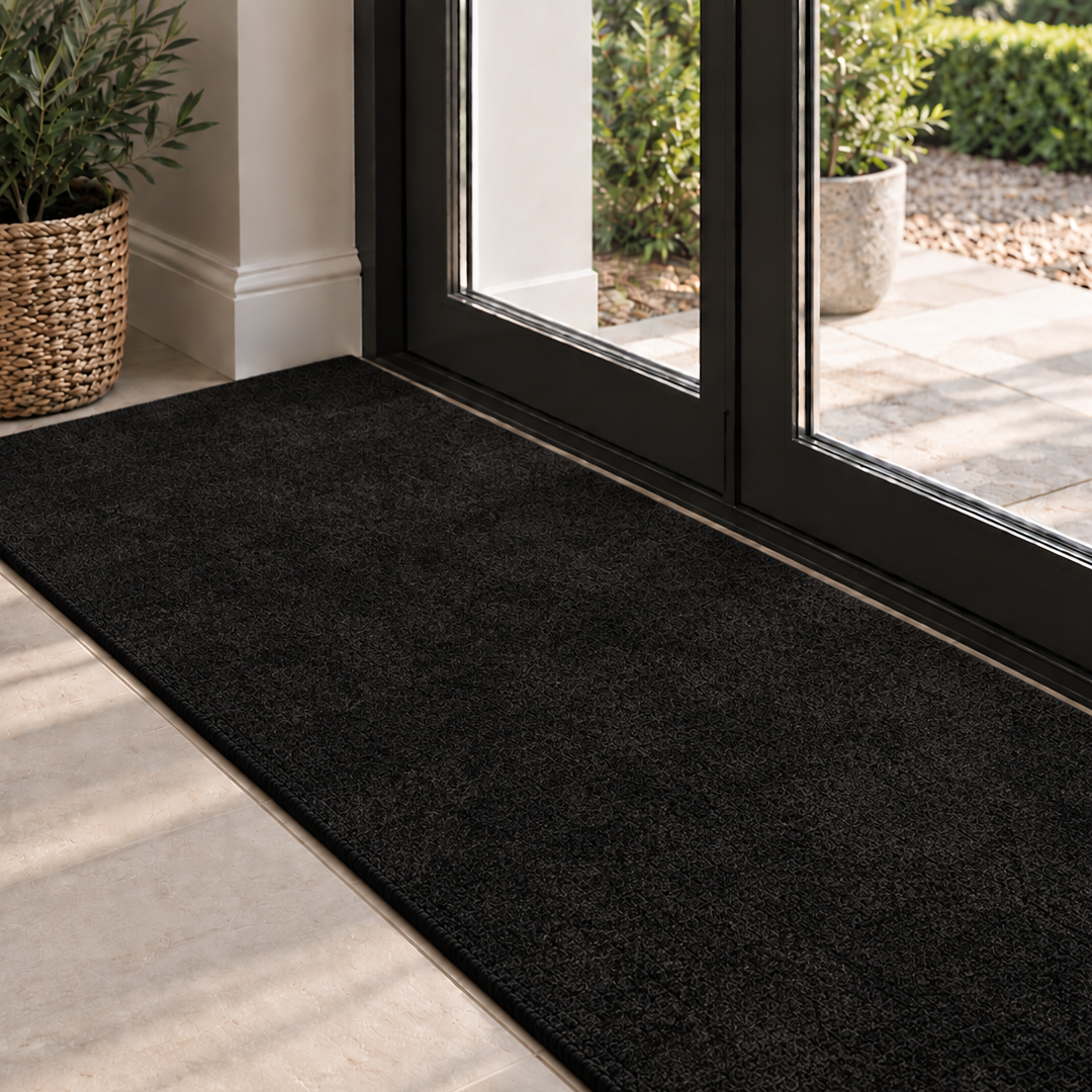 A long brownentryway doormat with a plush, high-pile texture, positioned on a light stone-tiled floor in front of black-framed glass double doors leading to a garden patio.